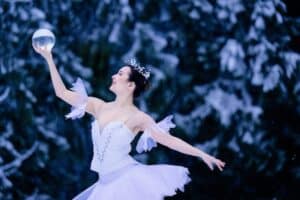 Ballerina in white tutu joyfully dances with a glass sphere in a snowy forest setting.