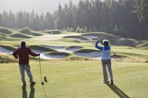 Senior woman golfer tees off with her male partner observing off to the side.